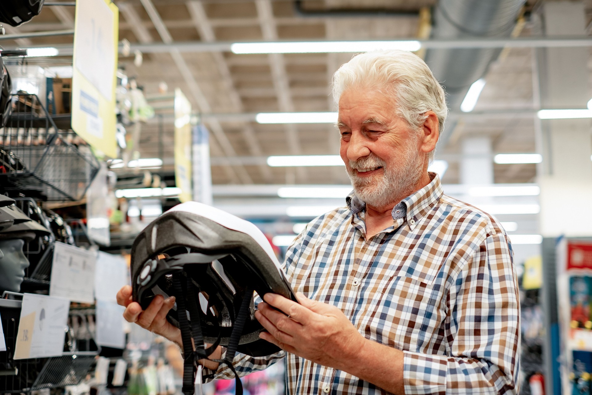 Elderly bearded man in shirt selects a bicycle protective helmet in a sports shop. He holds the helmet in hand, checking its technical indications. Consumerism concept